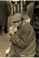 A man in a cap and worn tweed coat eats a sandwich as he sits on the base of a street lamp